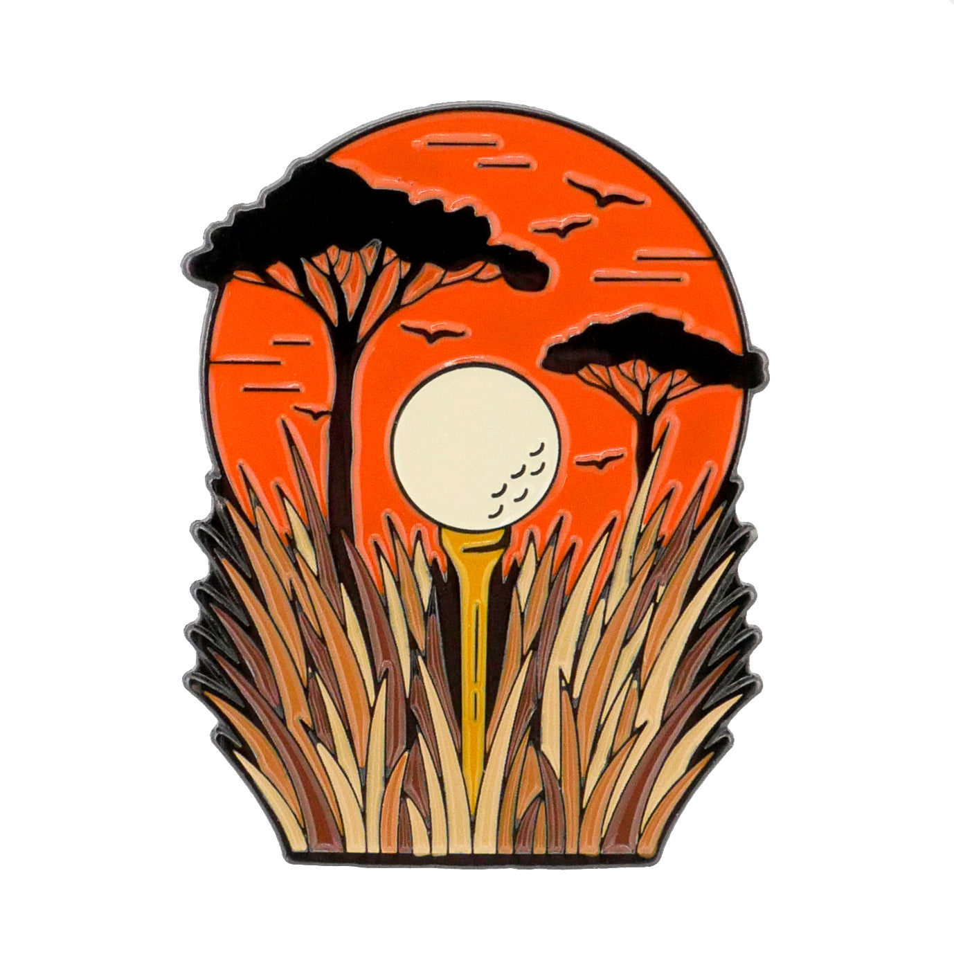 A golf ball on a tee stands in tall grass, framed by trees and an orange sunset sky with flying birds—a perfect scene, captured by the Sunset Tee Time Ball Marker from Ballmarkers.com.