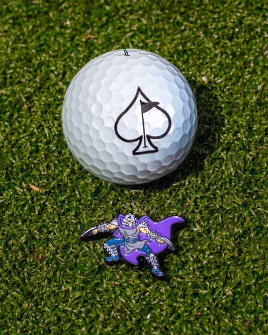 A golf ball with a black spade sits on green grass next to the TMNT - Shredder Ball Marker by Pins and Aces, featuring the cartoon armored villain from Teenage Mutant Ninja Turtles in purple and silver.