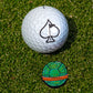 A white golf ball with a black spade symbol rests on green grass next to the Pins and Aces TMNT - Shell Shock Ball Marker, which is round and features green, orange, and black details.