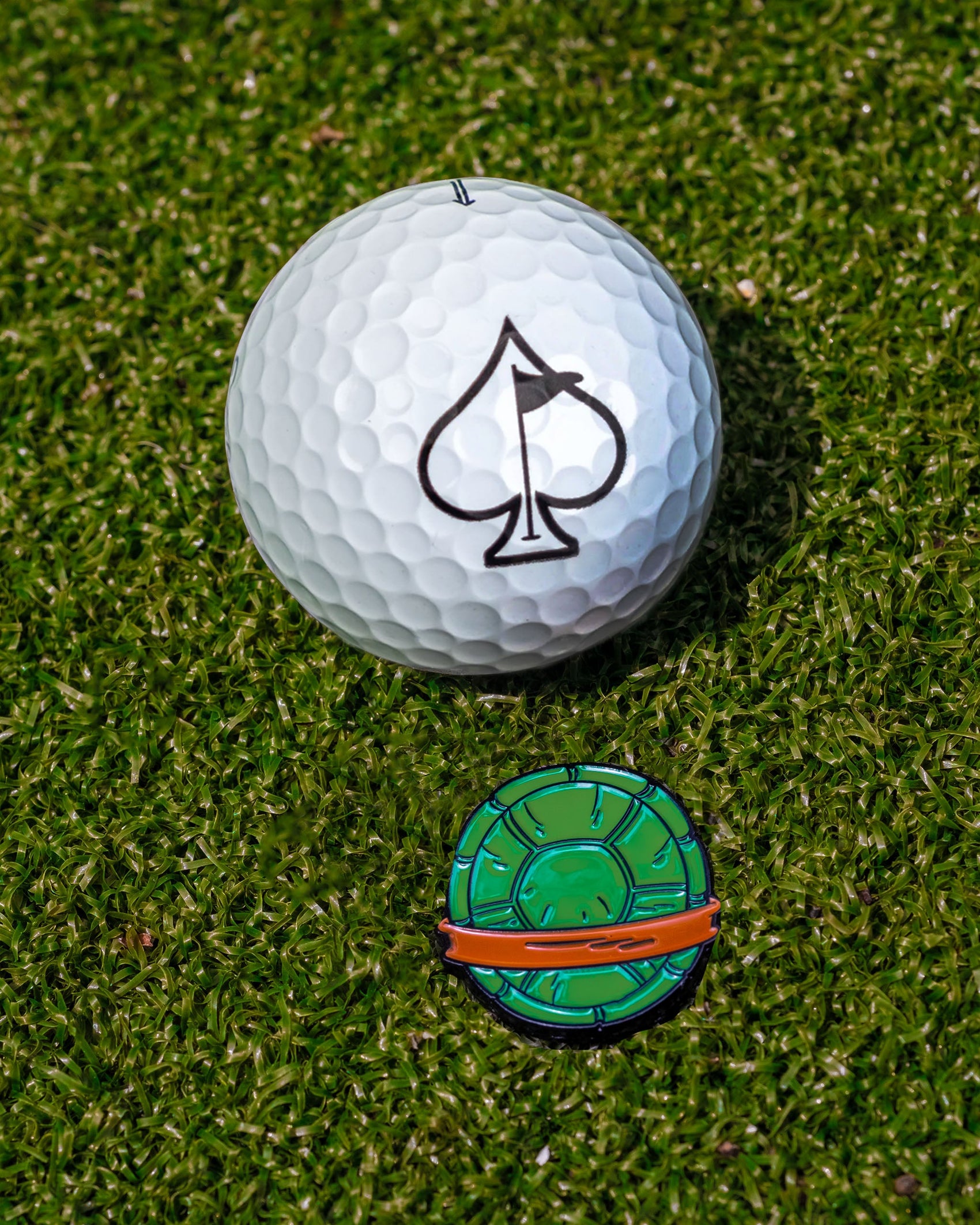 A white golf ball with a black spade symbol rests on green grass next to the Pins and Aces TMNT - Shell Shock Ball Marker, which is round and features green, orange, and black details.