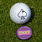 A white golf ball with an ace of spades logo sits on grass next to the TMNT - Sewer Cover Ball Marker by Pins and Aces, featuring a purple and pink geometric design with "IMNT" in yellow—perfect for Teenage Mutant Ninja Turtles fans.