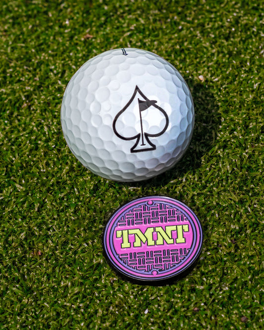A white golf ball with an ace of spades logo sits on grass next to the TMNT - Sewer Cover Ball Marker by Pins and Aces, featuring a purple and pink geometric design with "IMNT" in yellow—perfect for Teenage Mutant Ninja Turtles fans.