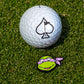 A white golf ball with an ace of spades logo sits on grass next to the Pins and Aces TMNT - Donatello Ball Marker— a playful Teenage Mutant Ninja Turtles accessory shaped like Donatello's green head with a purple mask.