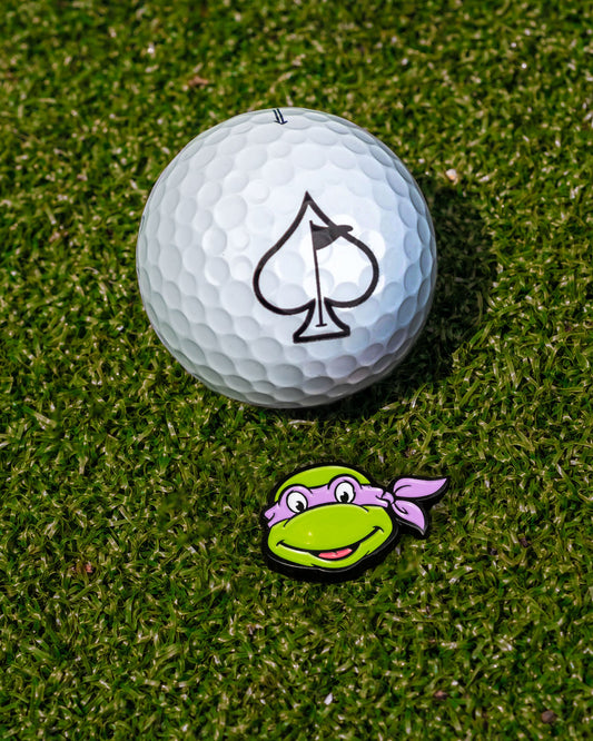 A white golf ball with an ace of spades logo sits on grass next to the Pins and Aces TMNT - Donatello Ball Marker— a playful Teenage Mutant Ninja Turtles accessory shaped like Donatello's green head with a purple mask.