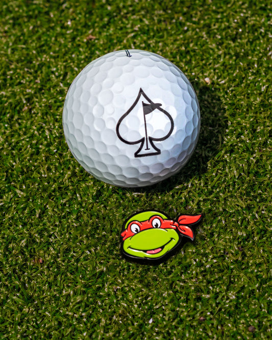A golf ball rests on green grass beside the Pins and Aces TMNT - Raphael Ball Marker, featuring a cartoon turtle with a red mask—the ideal accessory for Teenage Mutant Ninja Turtles fans.