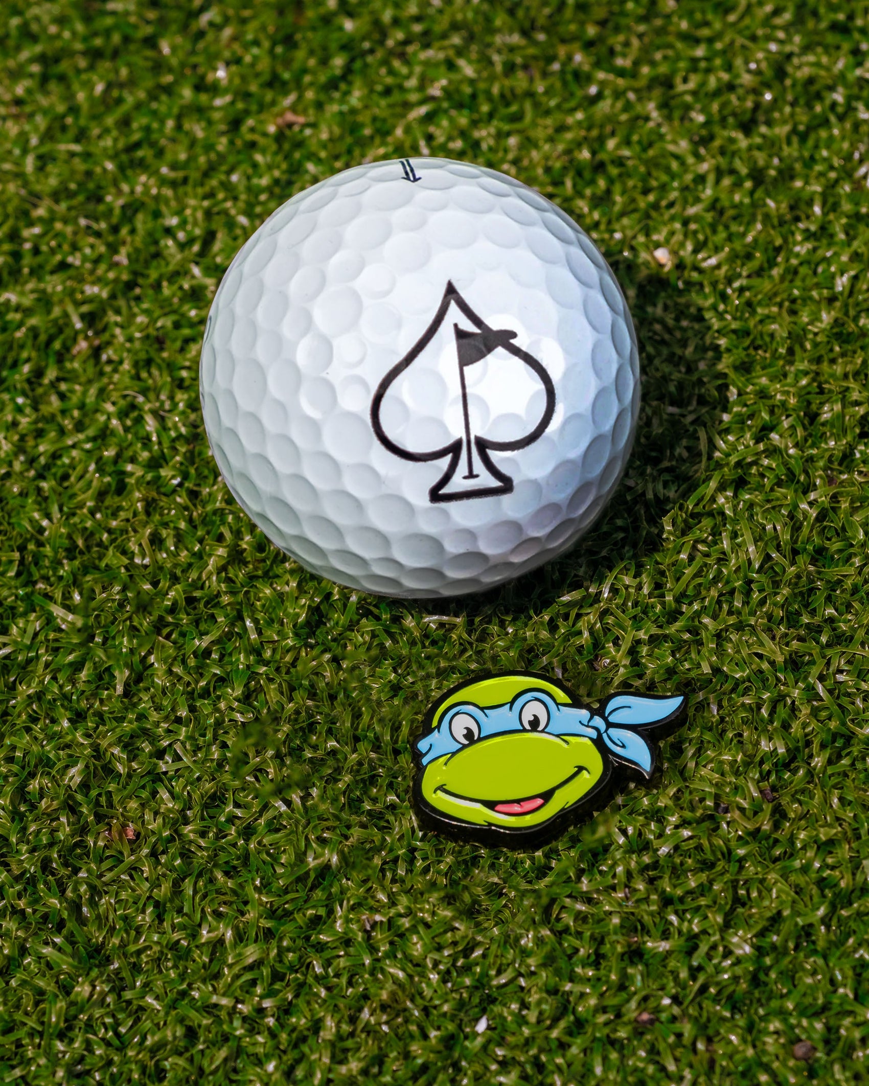 The Pins and Aces TMNT - Leonardo Ball Marker, featuring a fun cartoon design, sits next to a white golf ball with a black spade logo on green grass—perfect for Teenage Mutant Ninja Turtles golf fans.