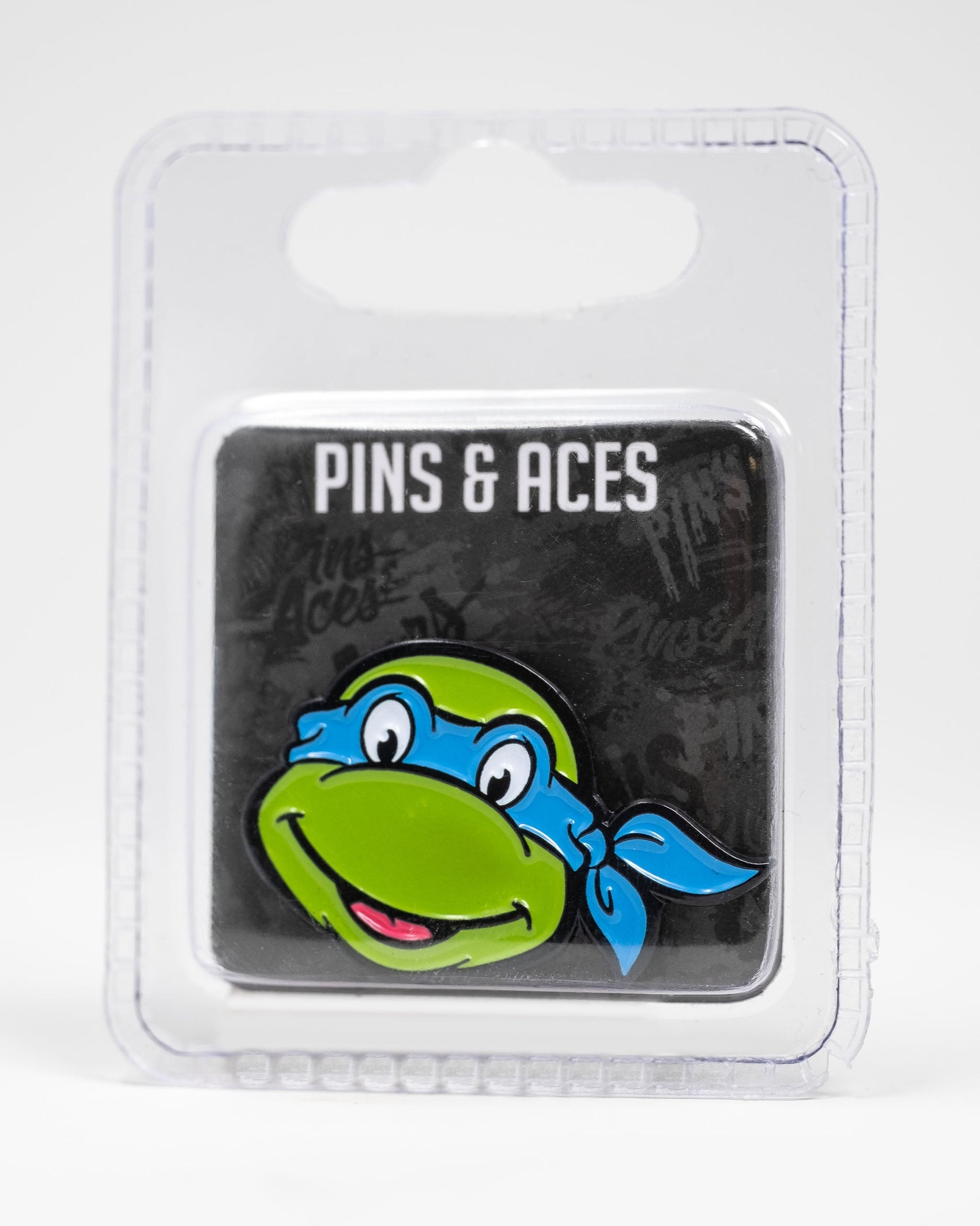 The Pins and Aces TMNT - Leonardo Ball Marker features a green cartoon turtle head with a blue mask and big smile, inspired by Leonardo, packaged in a clear case—an essential Teenage Mutant Ninja Turtles golf accessory.