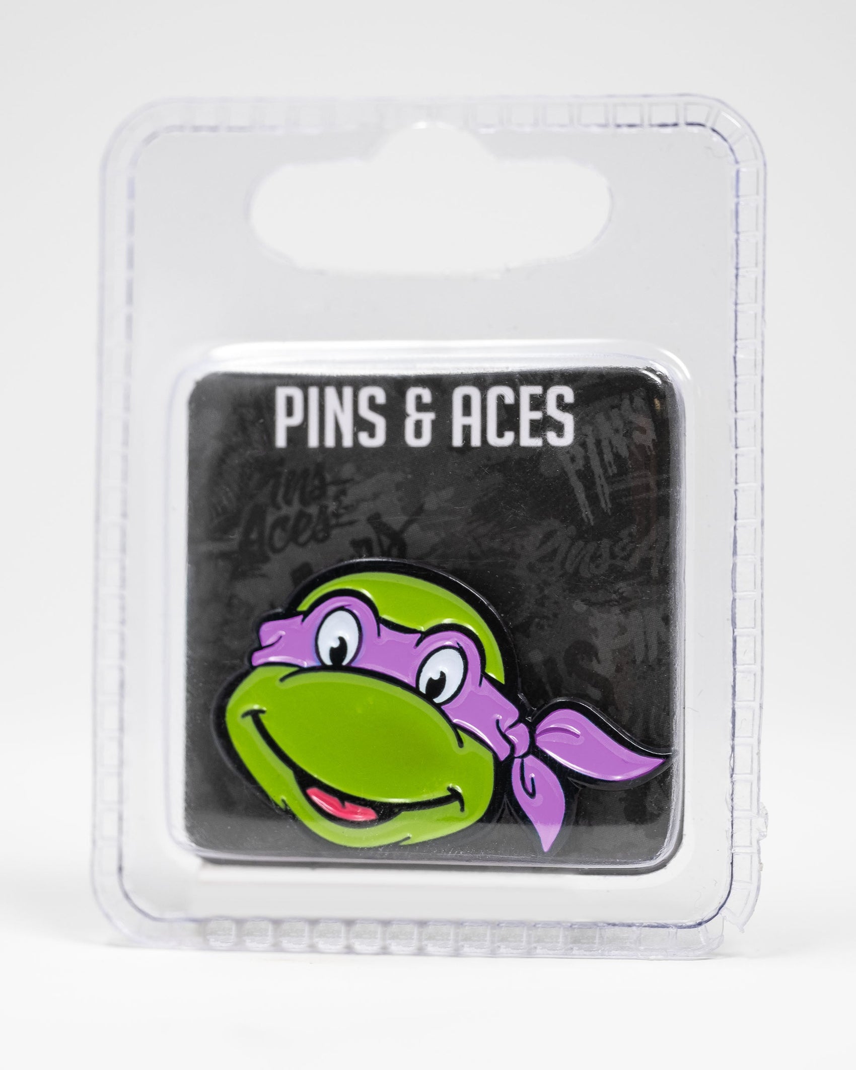 A TMNT - Donatello Ball Marker by Pins and Aces, featuring a cartoon turtle face with a purple mask, is shown on a black Pins and Aces card inside clear plastic packaging against a white background.