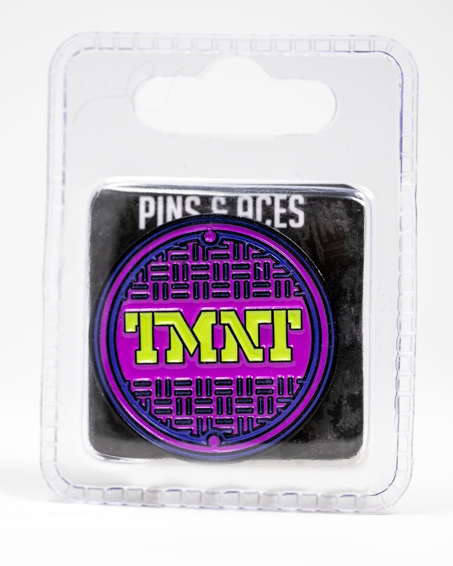 The TMNT - Sewer Cover Ball Marker by Pins and Aces is a round enamel pin featuring a purple and black sewer cover design with yellow TMNT letters, packaged in clear plastic labeled "Pins and Aces" at the top—ideal for Ninja Turtles golf fans.