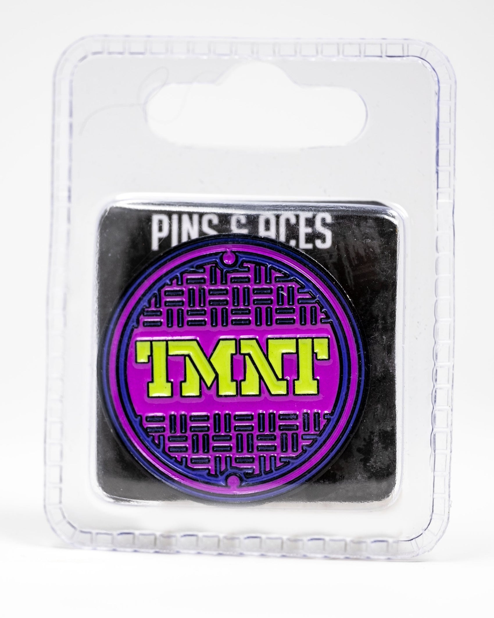 The TMNT - Sewer Cover Ball Marker by Pins and Aces is a round enamel pin featuring a purple and black sewer cover design with yellow TMNT letters, packaged in clear plastic labeled "Pins and Aces" at the top—ideal for Ninja Turtles golf fans.