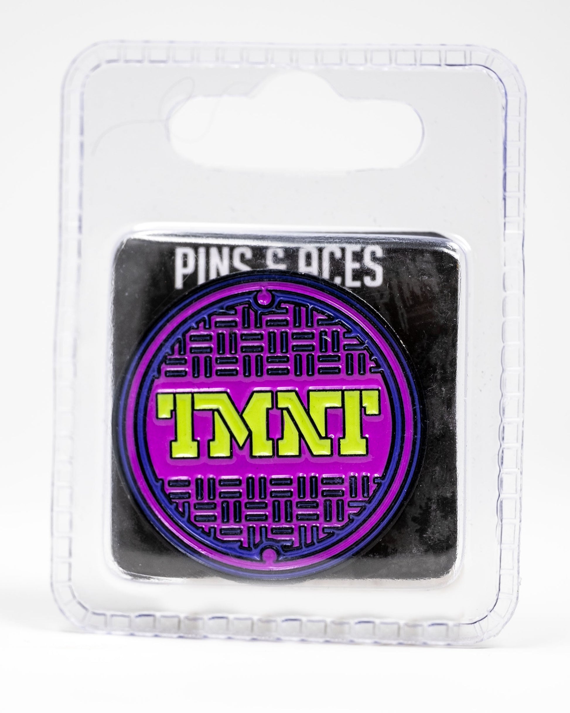 The TMNT - Sewer Cover Ball Marker by Pins and Aces is a round enamel pin featuring a purple and black sewer cover design with yellow TMNT letters, packaged in clear plastic labeled "Pins and Aces" at the top—ideal for Ninja Turtles golf fans.