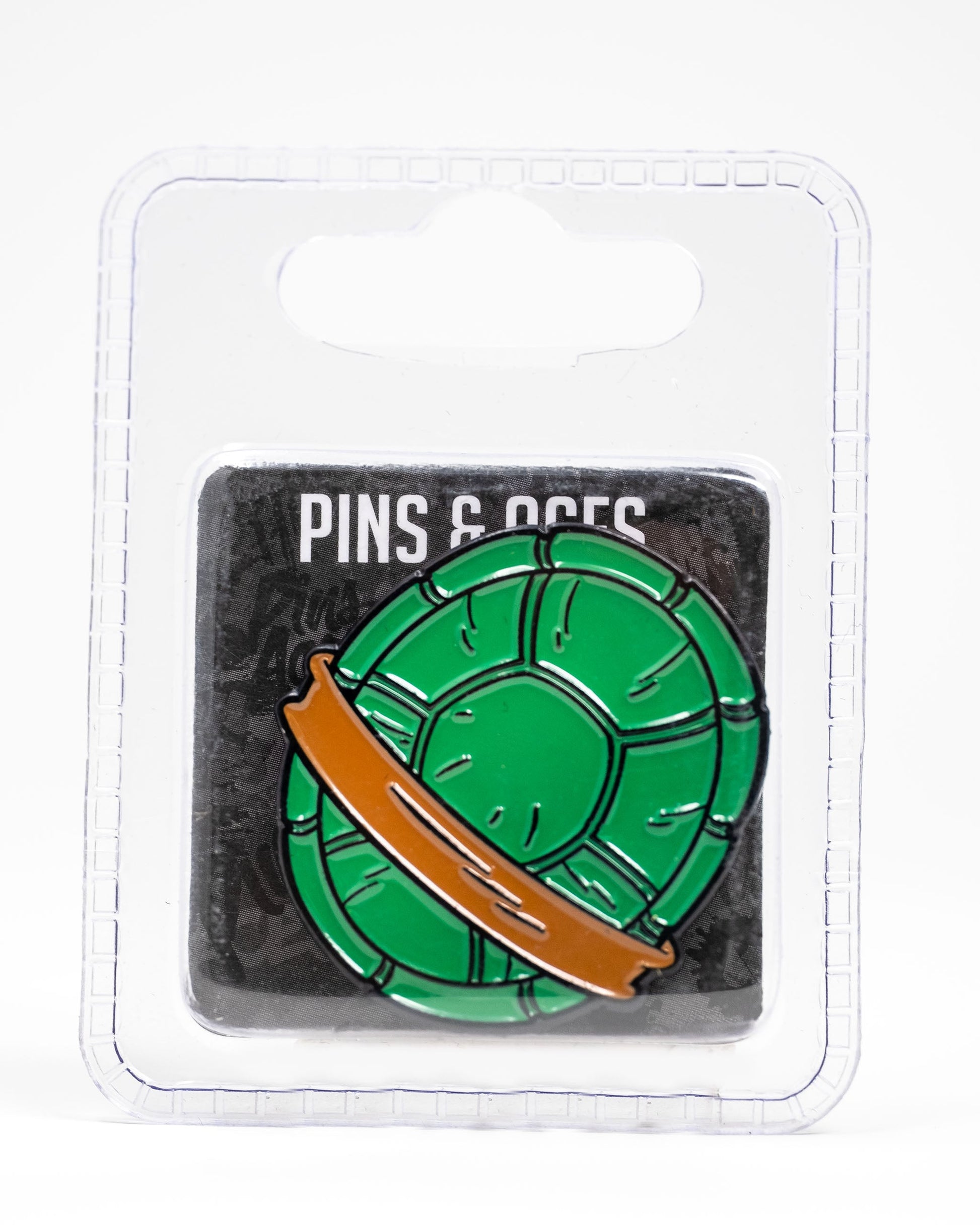 The TMNT - Shell Shock Ball Marker by Pins and Aces features a green enamel turtle shell with an orange band, inspired by Teenage Mutant Ninja Turtles, and comes on a branded card inside clear plastic packaging.