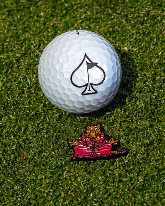 A golf ball with a black spade symbol rests on green grass beside the TMNT - Splinter Ball Marker by Pins and Aces, showcasing a meditating anthropomorphic tiger in red robes—a standout golf accessory.