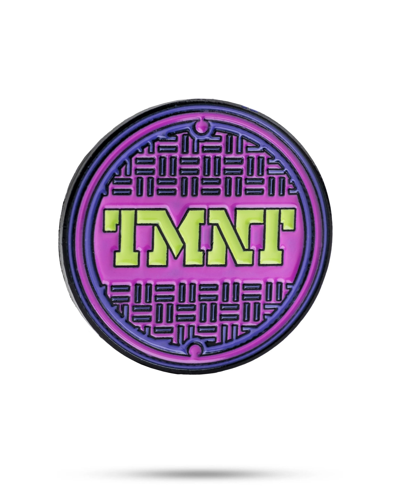 The TMNT - Sewer Cover Ball Marker by Pins and Aces is a round pin with a purple and black manhole cover design, inspired by TMNT, featuring bold neon green letters at the center. Isolated on a white background with a slight shadow.