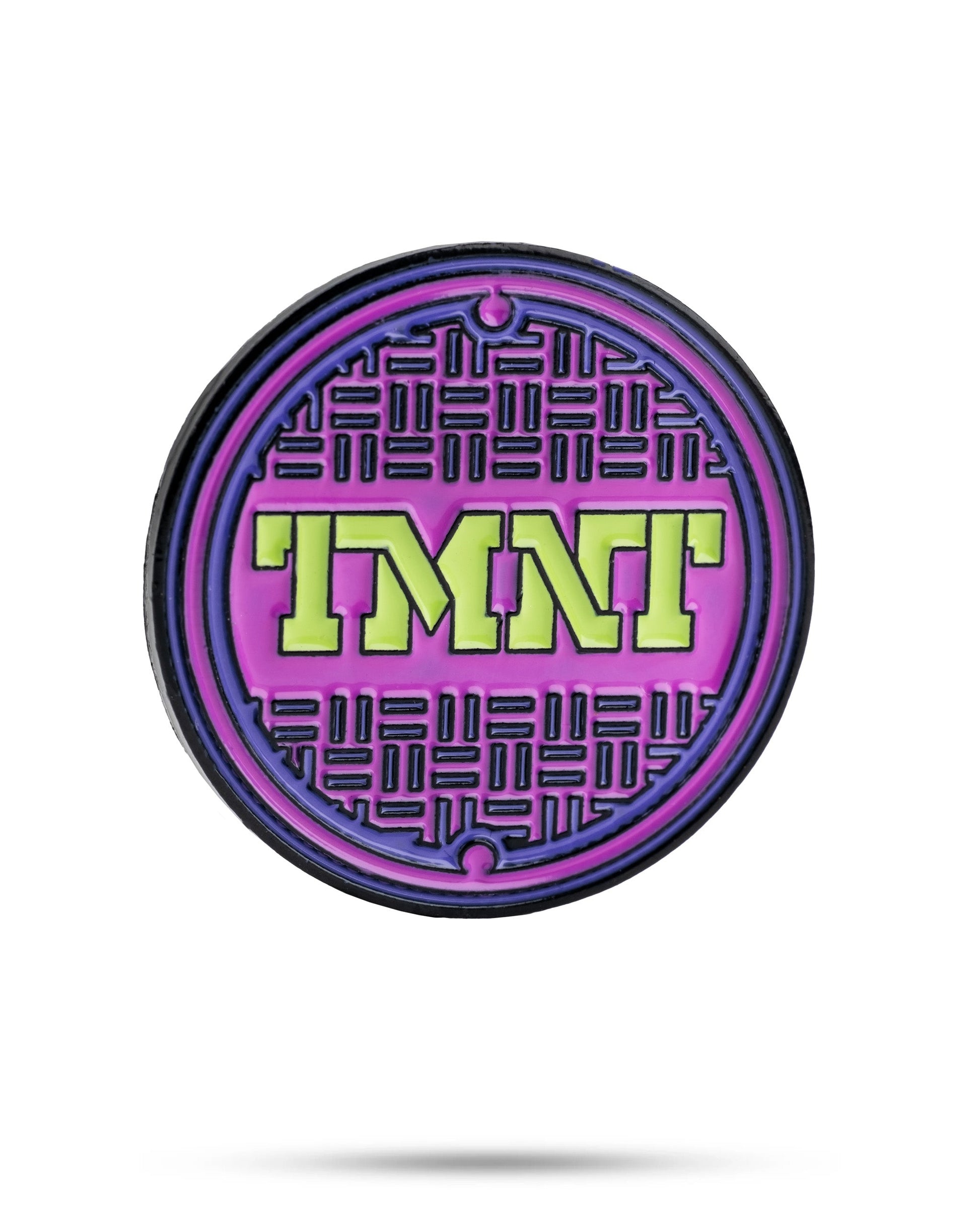 The TMNT - Sewer Cover Ball Marker by Pins and Aces is a round pin with a purple and black manhole cover design, inspired by TMNT, featuring bold neon green letters at the center. Isolated on a white background with a slight shadow.