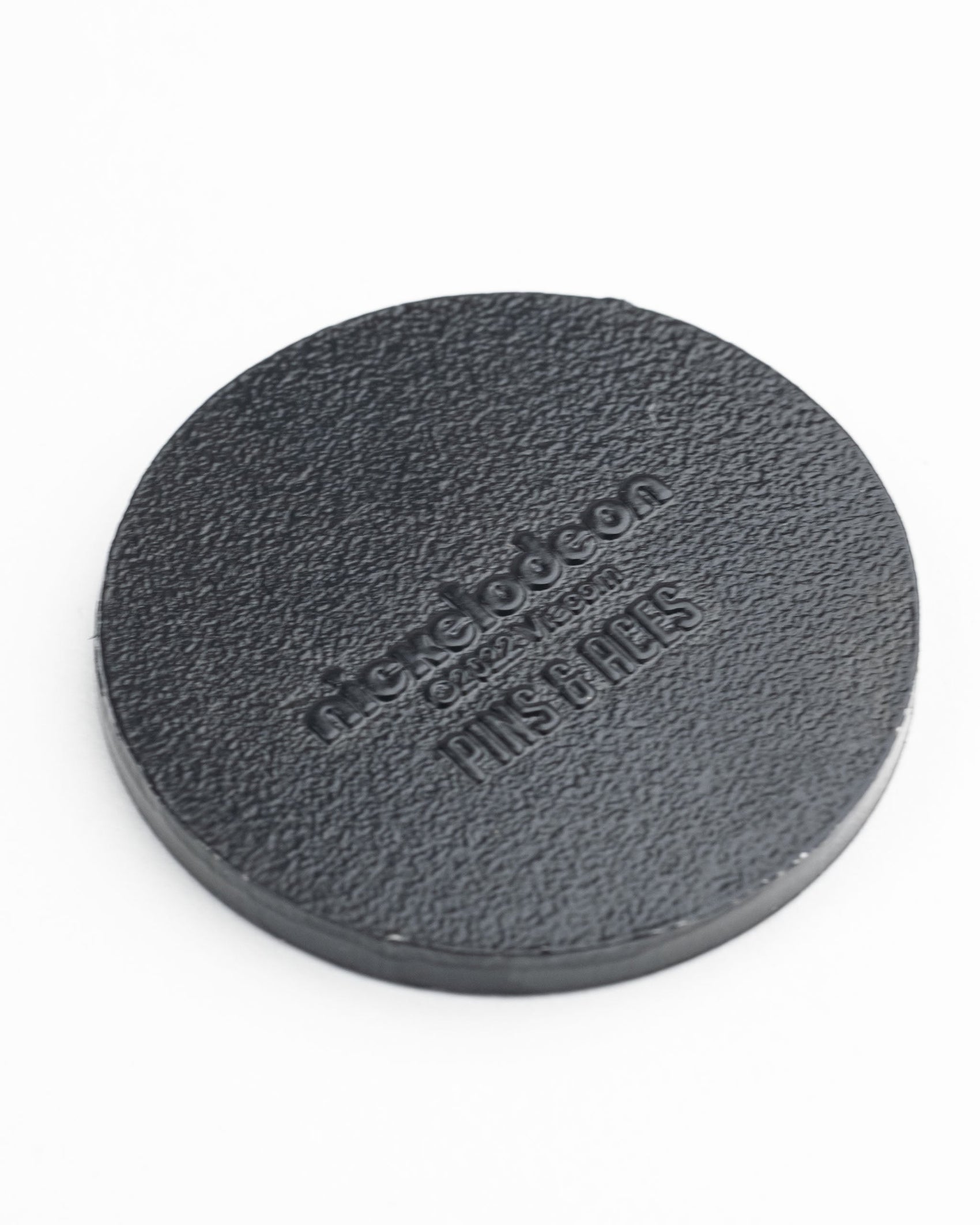 The TMNT - Sewer Cover Ball Marker by Pins and Aces is a round, textured black marker resembling a sewer cover, embossed with Nickelodeon and Pins & Tags, shown on a white background.