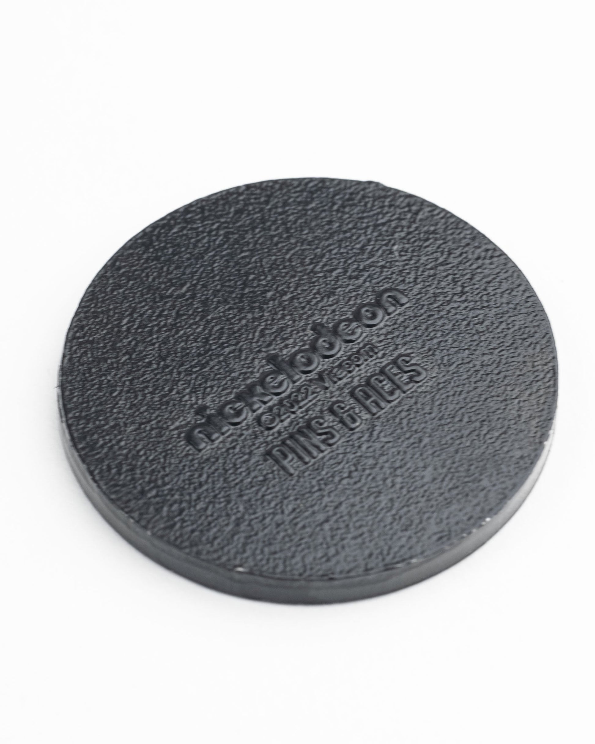 The TMNT - Sewer Cover Ball Marker by Pins and Aces is a round, textured black marker resembling a sewer cover, embossed with Nickelodeon and Pins & Tags, shown on a white background.