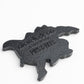 A black plastic TMNT - Shredder Ball Marker by Pins and Aces, turtle-shaped with raised “nextdoossn” and “PLUS OF AGES” text, resembling a Teenage Mutant Ninja Turtles golf accessory, shown on a white background.
