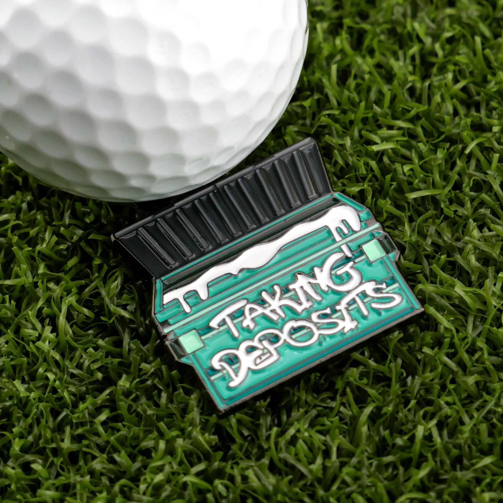 A golf ball sits on green grass next to Logie Joe's Golf’s "Taking Deposits" marker, shaped like a green dumpster with white text and a snow-like line—one of the funniest premium golf accessories around.