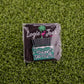 A square pin on grass shows Logie Joe’s Golf’s "Taking Deposits"—a palm tree, flamingo, and green dumpster with white liquid overflowing—making it a hilarious ball marker or fun golf accessory.