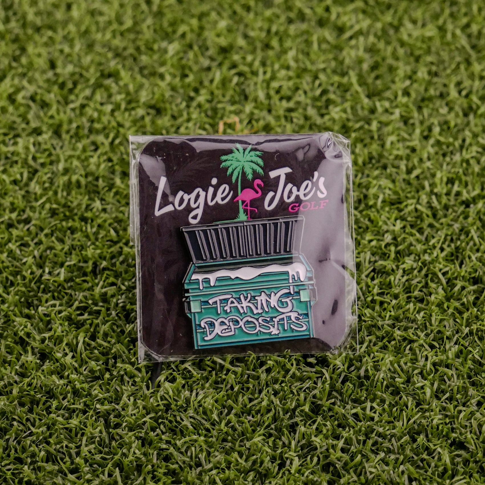 A square pin on grass shows Logie Joe’s Golf’s "Taking Deposits"—a palm tree, flamingo, and green dumpster with white liquid overflowing—making it a hilarious ball marker or fun golf accessory.