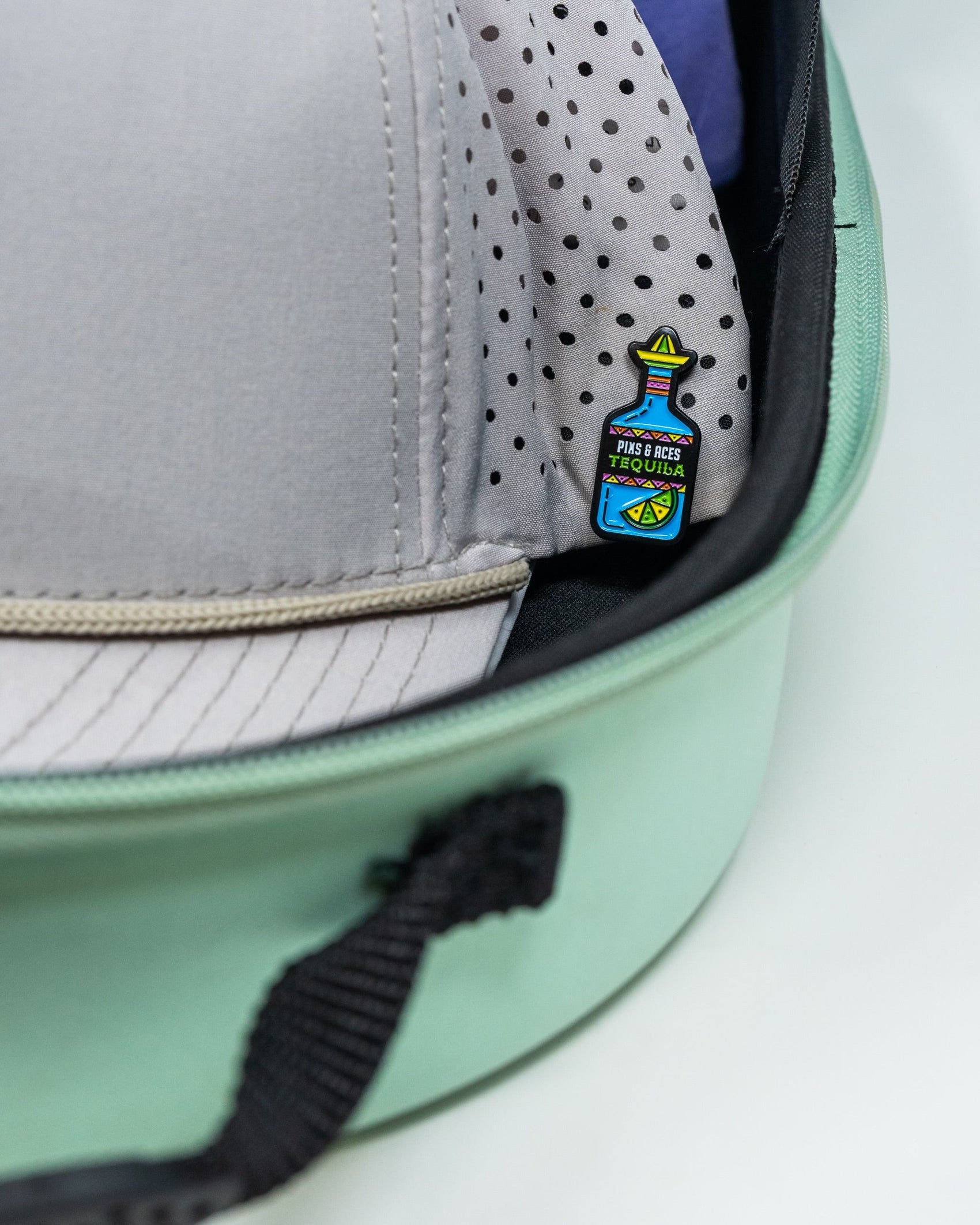 A light grey cap with a colorful tequila bottle pin is stored in a Pins and Aces Hat Caddie, featuring a mint green exterior, black zipper, and polka dot lining.
