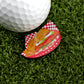 A golf ball on artificial grass sits beside a premium Logie Joe's Golf ball marker, featuring a funny Breaded Tenderloin Sandwich Basket design with fries on a checkered plate.