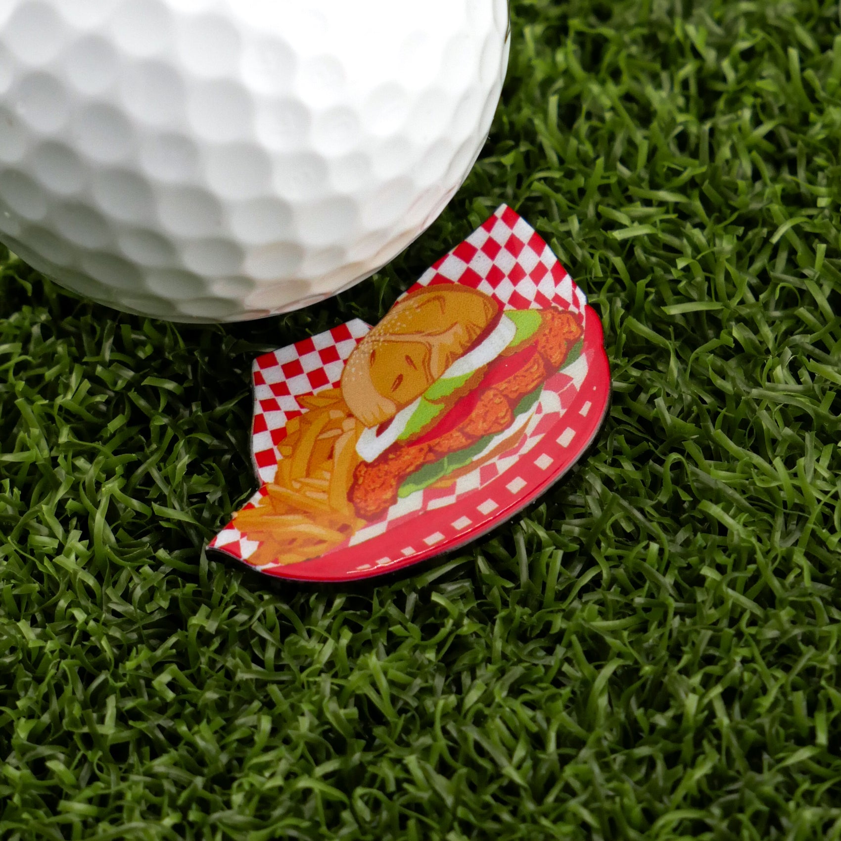 A golf ball on artificial grass sits beside a premium Logie Joe's Golf ball marker, featuring a funny Breaded Tenderloin Sandwich Basket design with fries on a checkered plate.