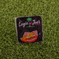 A square sticker featuring Logie Joe's Golf, a palm tree and flamingo logo, and the Breaded Tenderloin Sandwich Basket sits on artificial green grass—perfect for fans of funny golf gear or quirky golf ball markers.