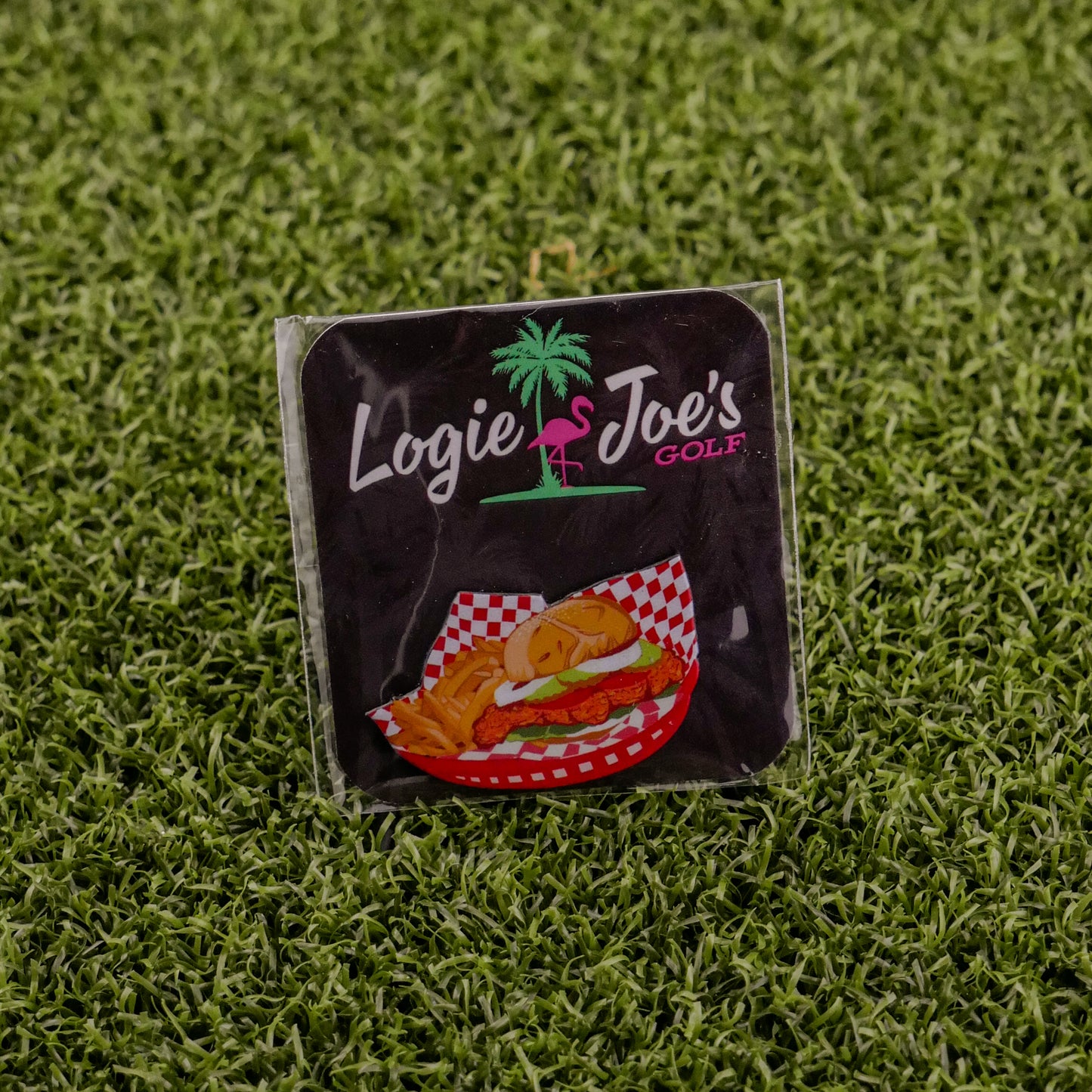 A square sticker featuring Logie Joe's Golf, a palm tree and flamingo logo, and the Breaded Tenderloin Sandwich Basket sits on artificial green grass—perfect for fans of funny golf gear or quirky golf ball markers.