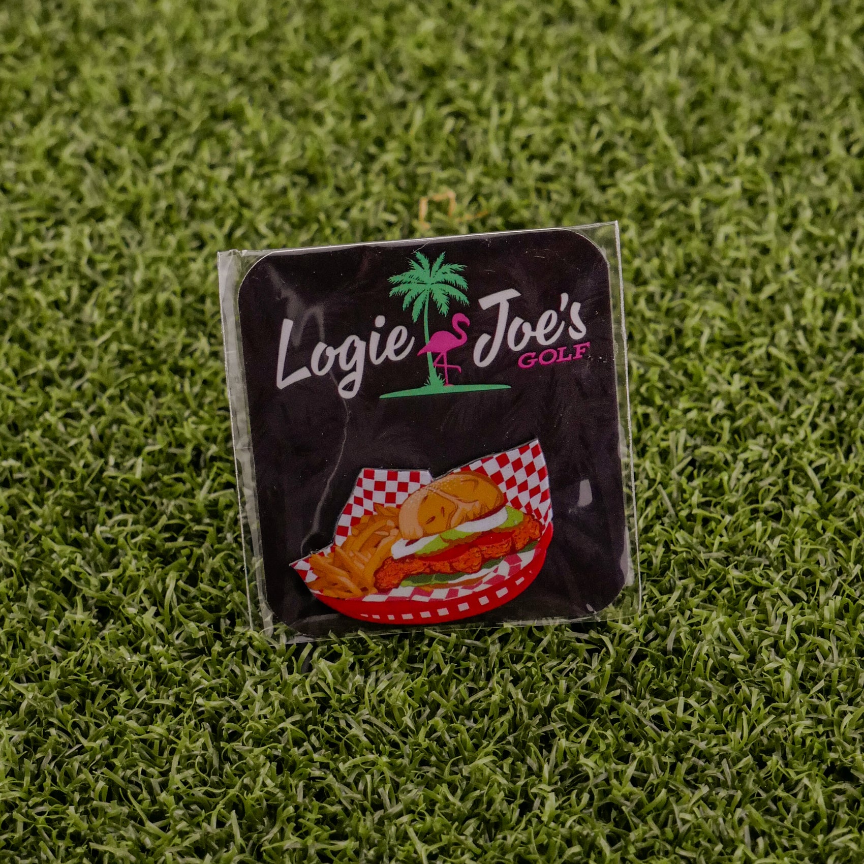 A square sticker featuring Logie Joe's Golf, a palm tree and flamingo logo, and the Breaded Tenderloin Sandwich Basket sits on artificial green grass—perfect for fans of funny golf gear or quirky golf ball markers.