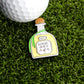 A golf ball rests on artificial grass beside the Tequila Ball Marker by Logie Joe's Golf, shaped like a bottle and featuring a small flamingo illustration—a unique accessory for a fun day on the course.