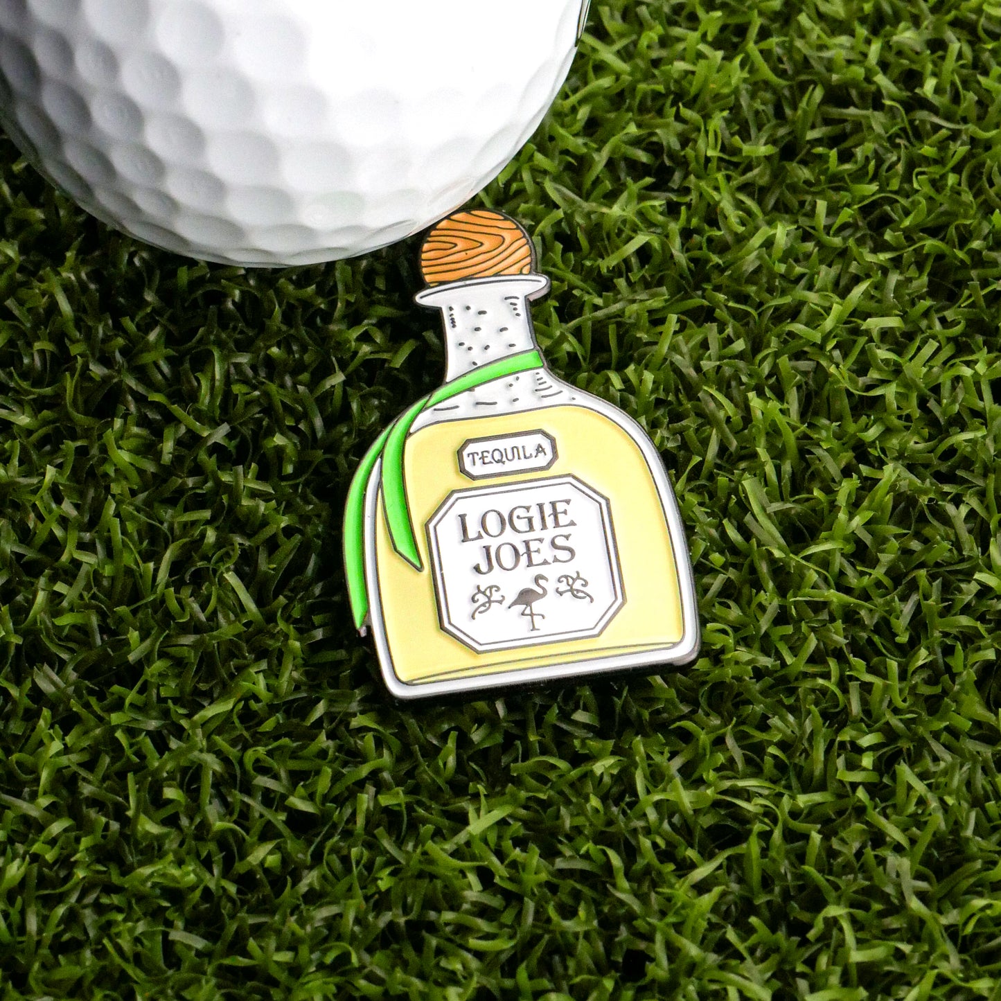 A golf ball rests on artificial grass beside the Tequila Ball Marker by Logie Joe's Golf, shaped like a bottle and featuring a small flamingo illustration—a unique accessory for a fun day on the course.
