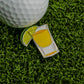 A golf ball rests on green artificial grass beside Logie Joe's Golf Tequila & Lime Ball Marker—an enamel pin shaped like a yellow drink with a lime wedge—for one of the funniest golf accessories on the course.
