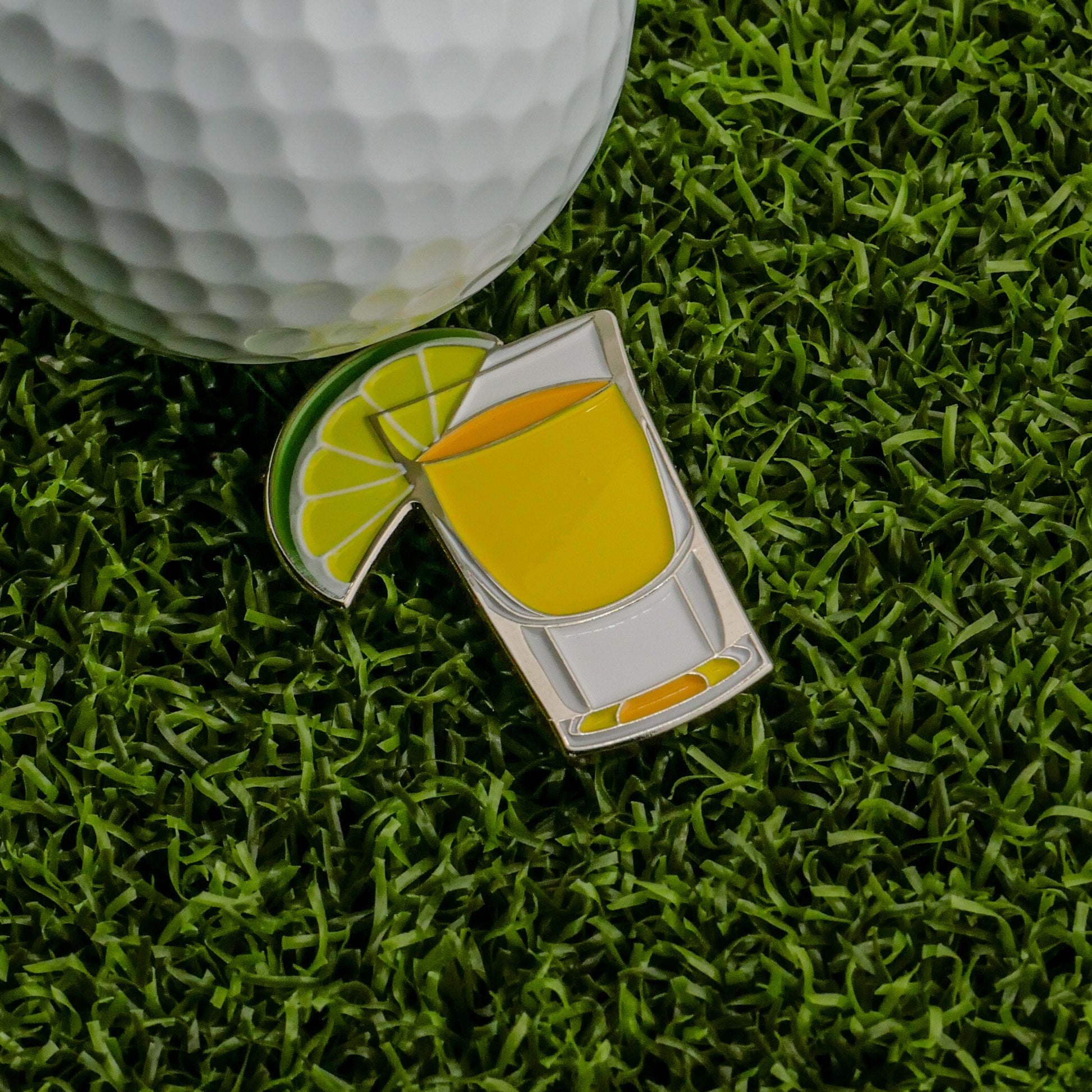 A golf ball rests on green artificial grass beside Logie Joe's Golf Tequila & Lime Ball Marker—an enamel pin shaped like a yellow drink with a lime wedge—for one of the funniest golf accessories on the course.