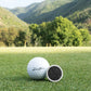A Doubloon Venom golf ball and a small round black marker sit on green grass with mountains, trees, and blue sky in the background—ideal for highlighting lightweight Titanium or forged Carbon Fiber gear.