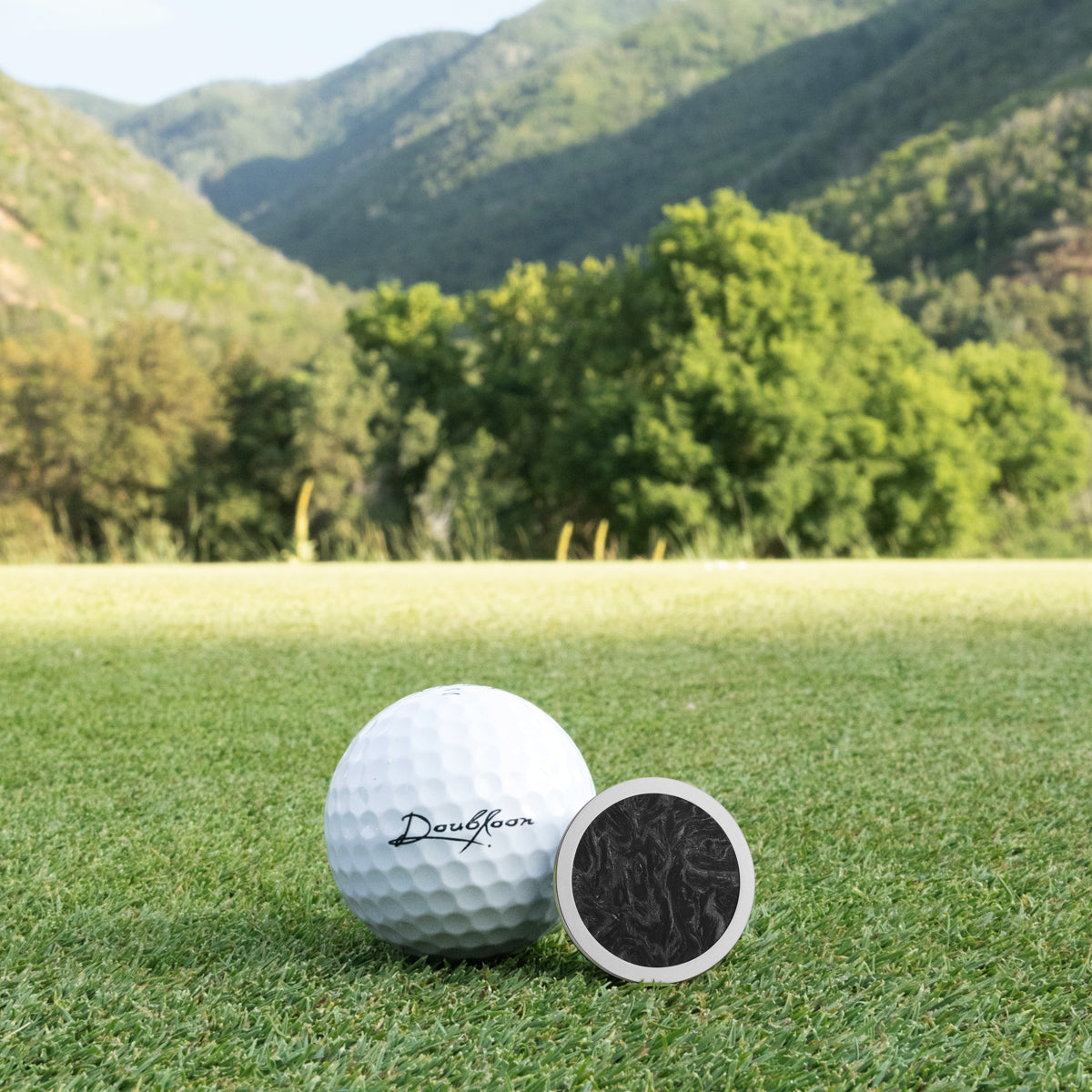 A Doubloon Venom golf ball and a small round black marker sit on green grass with mountains, trees, and blue sky in the background—ideal for highlighting lightweight Titanium or forged Carbon Fiber gear.