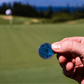 A hand holds The Venom by Doubloon, a blue golf ball marker with a titanium base, on a golf course with a softly blurred green, flag, and trees in the background.