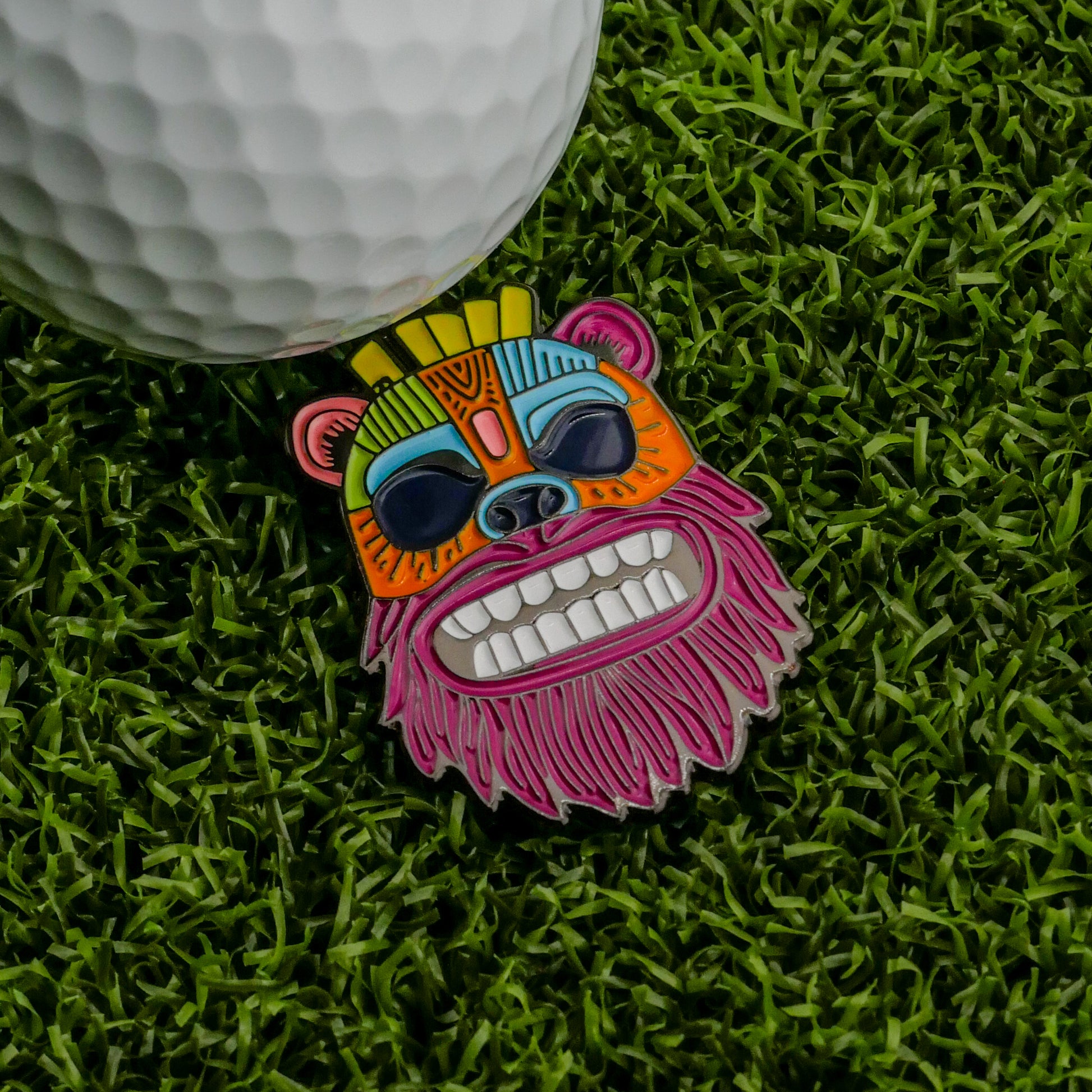 A vibrant Tiki Bear Ball Marker from Logie Joe's Golf, featuring a playful bear design with sunglasses and big teeth, sits on artificial grass beside a white golf ball—one of the most unique markers at Ballmarkers.com.