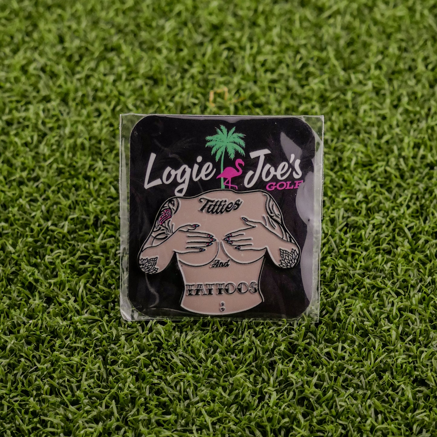 The Titties & Tattoos premium square ball marker by Logie Joe's Golf features a topless tattooed torso, pink flamingo, and palm tree on artificial grass—perfect for those who love funny golf accessories.