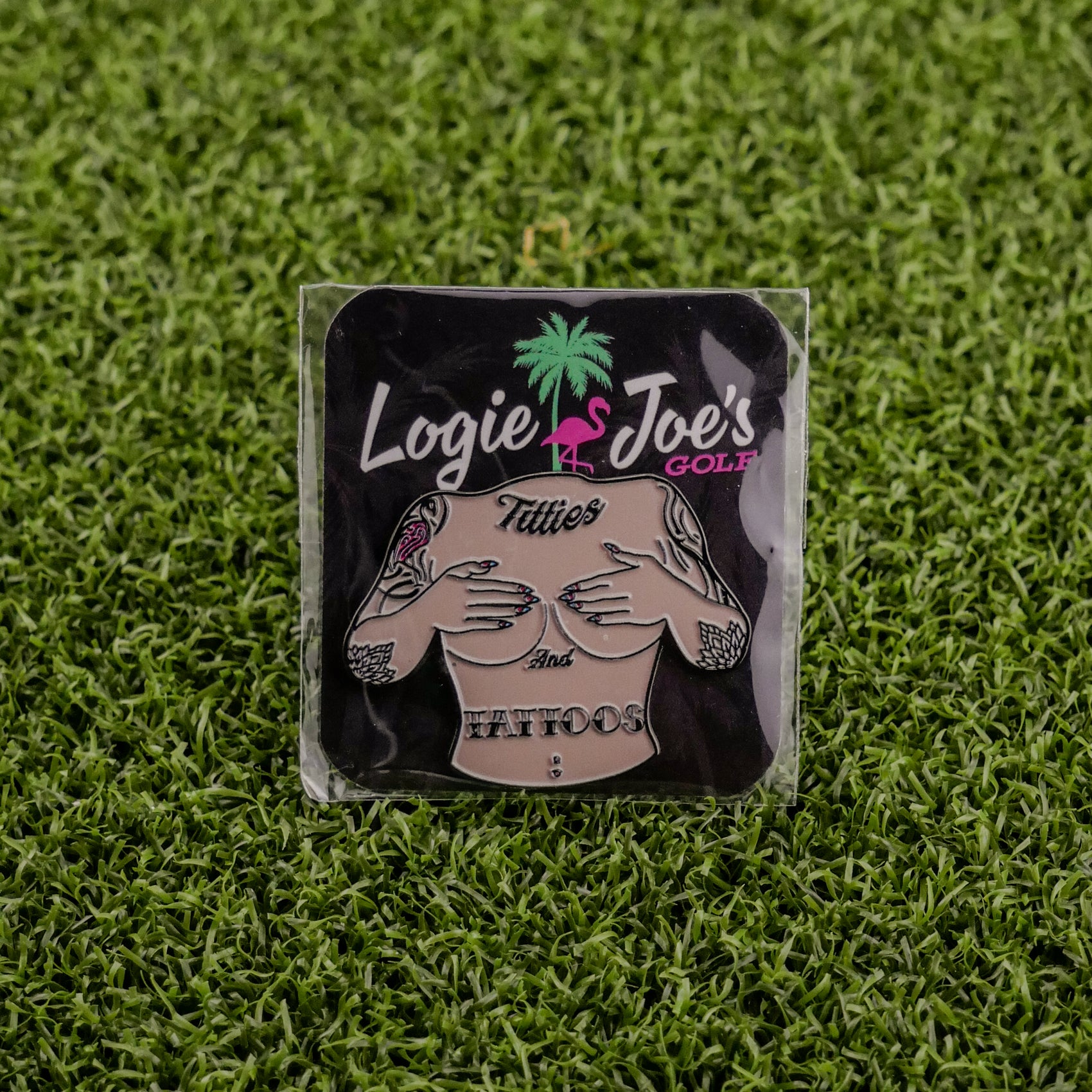 The Titties & Tattoos premium square ball marker by Logie Joe's Golf features a topless tattooed torso, pink flamingo, and palm tree on artificial grass—perfect for those who love funny golf accessories.