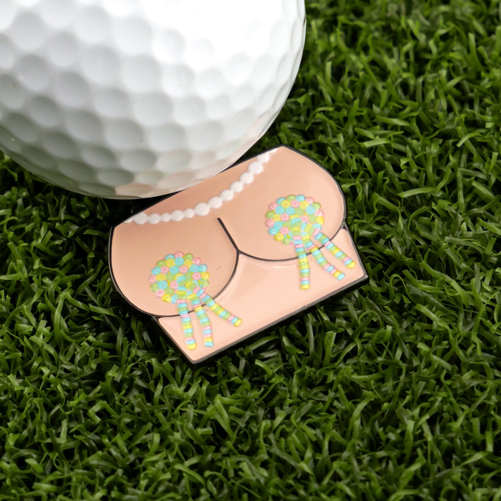 Logie Joe's Golf Titty Tassles is a hilarious golf ball marker shaped like cartoon buttocks with tassels, perfect for golf lovers who enjoy a good laugh on the course. Displayed here next to a golf ball on artificial grass.