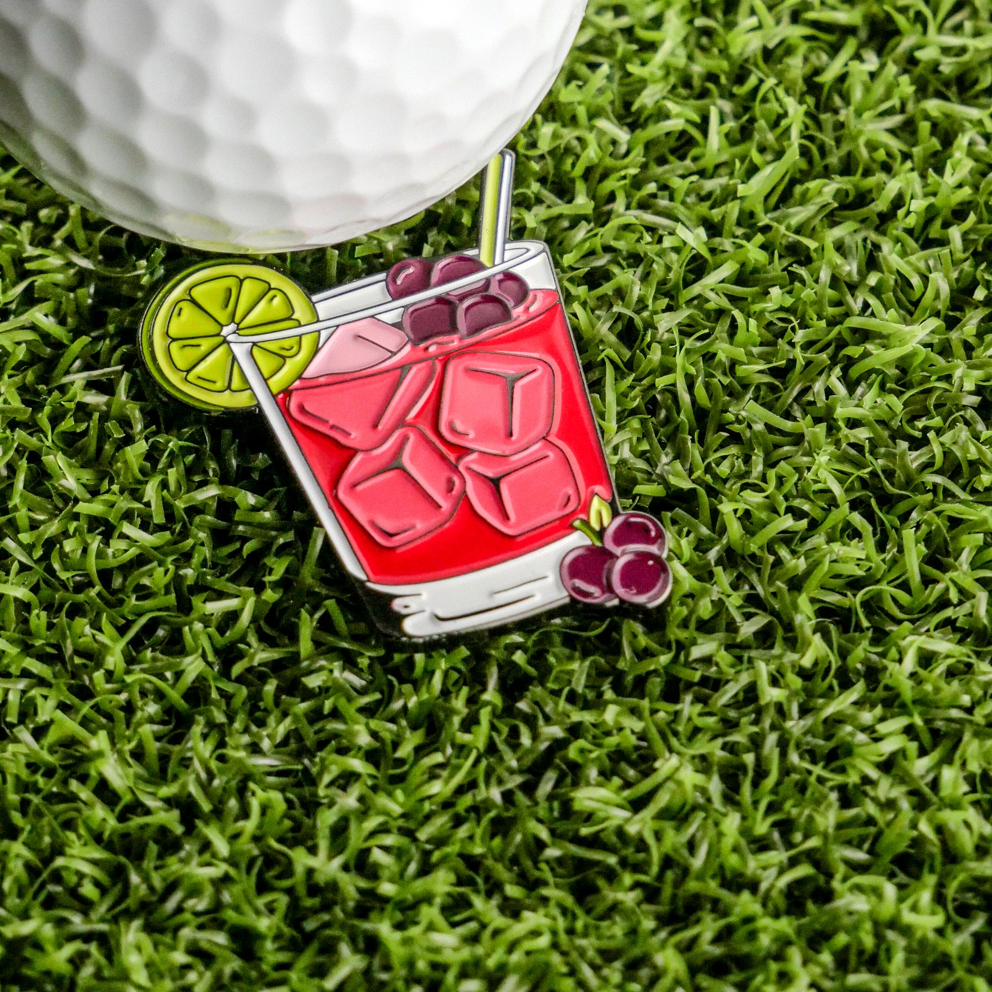 A golf ball sits on artificial grass next to the Transfusion Ball Marker from Ballmarkers.com, styled as a red iced drink with lime, straw, ice cubes, and cherries—an ideal addition to your cocktail collection.