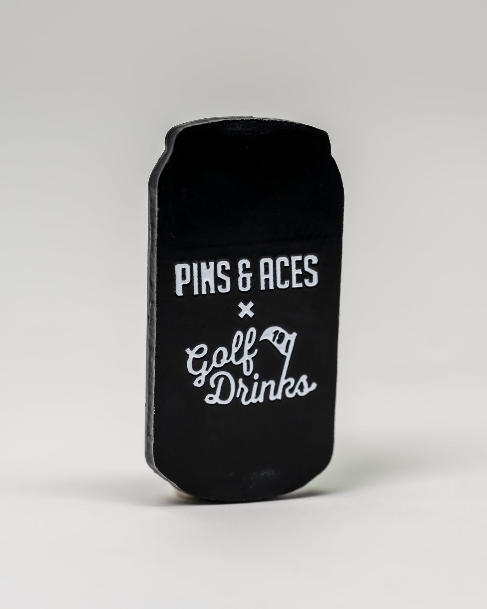 A black, soda can-shaped Pins and Aces Transfusion Can Ball Marker features white "PINS & ACES x Golf Drinks" text, a small golf club and ball graphic, and is designed to hold magnetic golf accessories or an enamel ball marker. Plain light background.