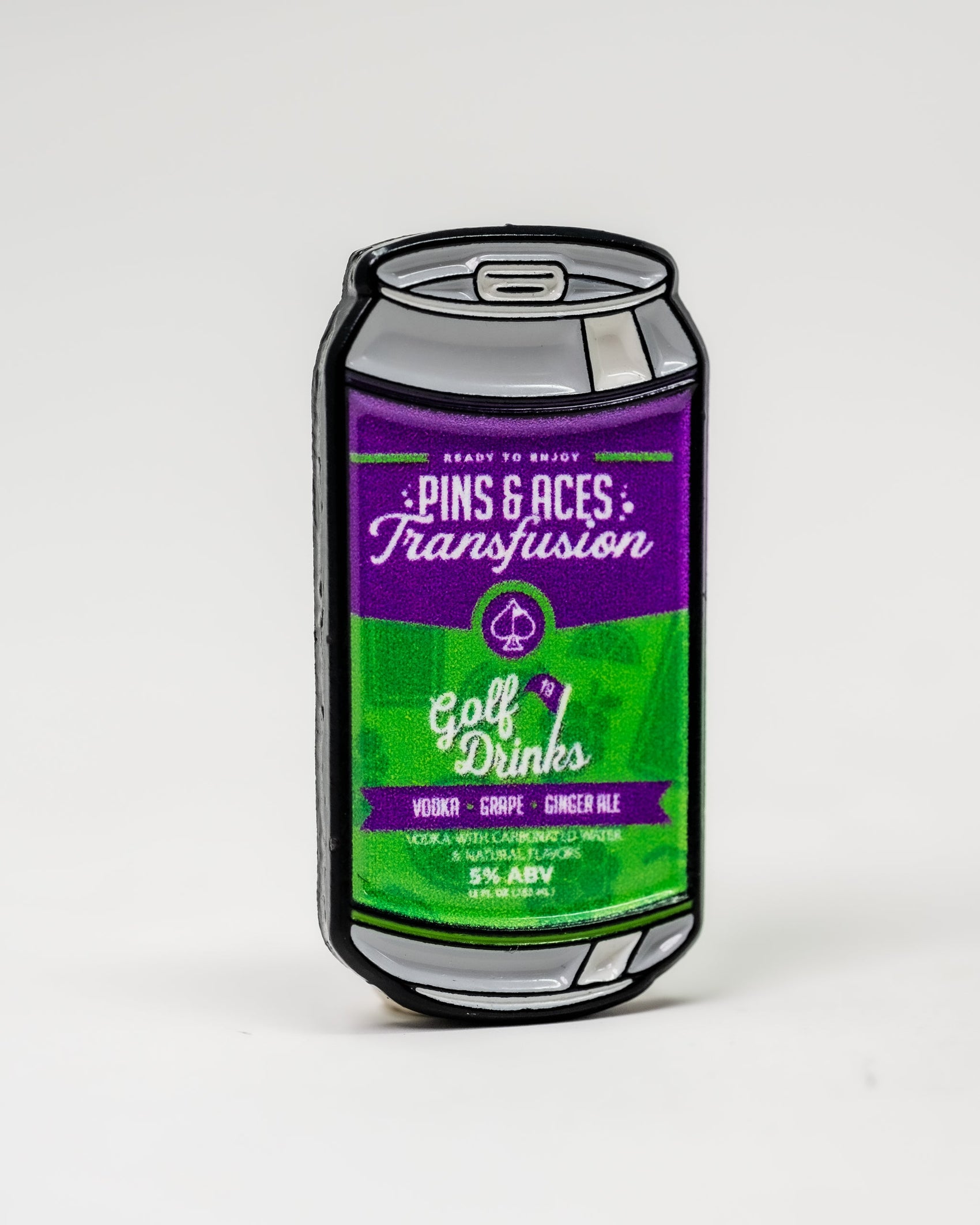 A Pins and Aces Transfusion Can Ball Marker is shown with a purple and green label reading “Transfusion,” featuring vodka, grape, ginger ale (5% ABV), and paired enamel magnetic golf accessories on a plain white background.