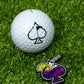 A white golf ball with a black transfusion spade logo sits on green grass next to the Pins and Aces Transfusion Spade Ball Marker, designed like a vibrant purple cocktail complete with a straw and lemon slice.