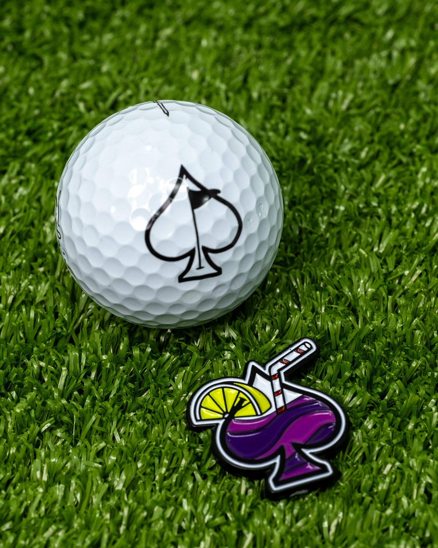 A white golf ball with a black transfusion spade logo sits on green grass next to the Pins and Aces Transfusion Spade Ball Marker, designed like a vibrant purple cocktail complete with a straw and lemon slice.