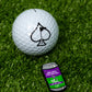 The Pins and Aces Transfusion Can Ball Marker, featuring a green and purple label, sits on artificial grass beside a golf ball with a large spade symbol—ideal for pairing with enamel markers or magnetic golf accessories.