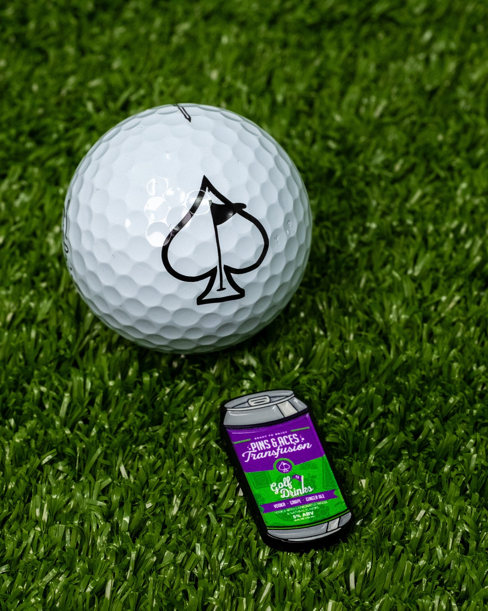 The Pins and Aces Transfusion Can Ball Marker, featuring a green and purple label, sits on artificial grass beside a golf ball with a large spade symbol—ideal for pairing with enamel markers or magnetic golf accessories.