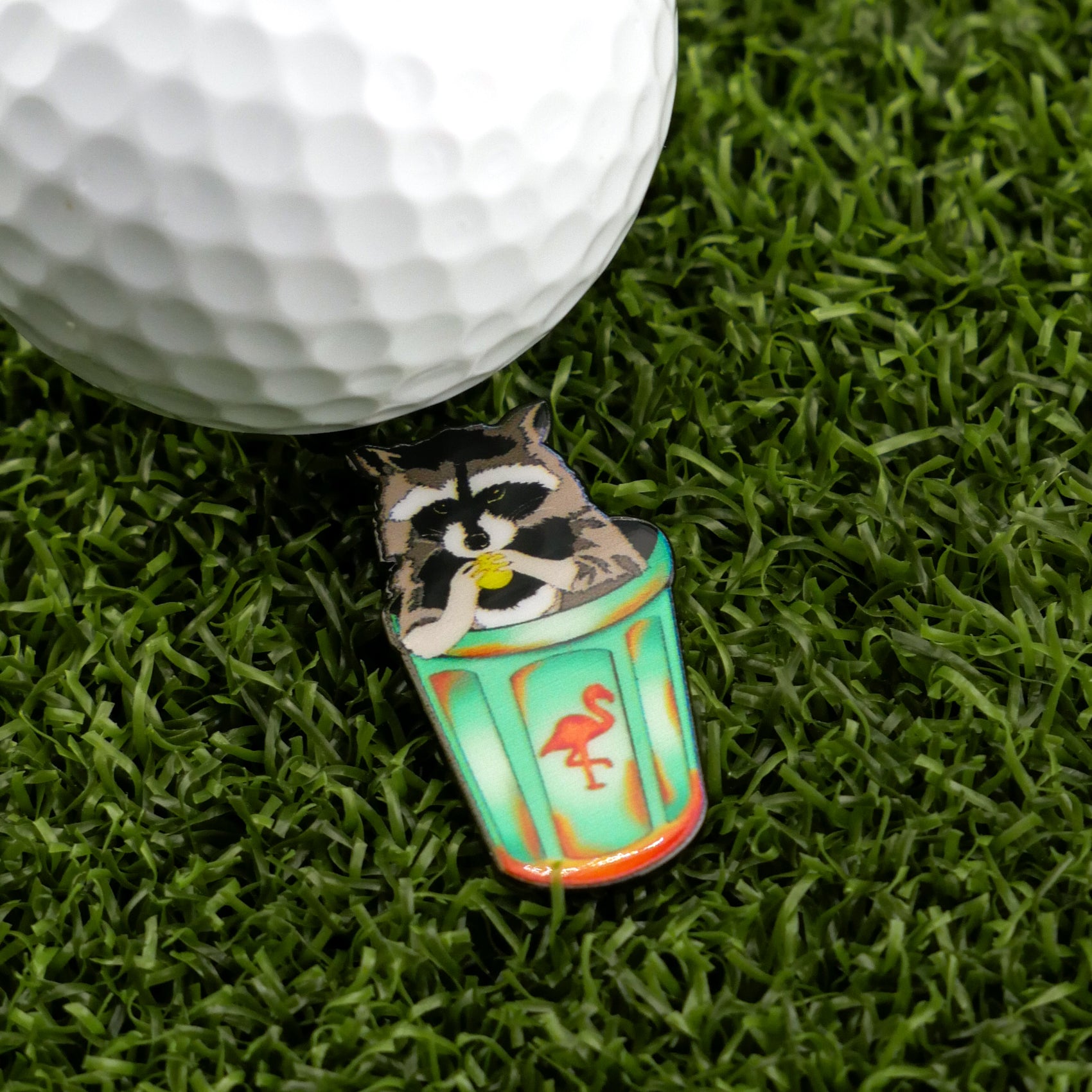 A golf ball sits on artificial grass beside the Trash Panda divot tool by Logie Joe's Golf, featuring a playful raccoon with a pink flamingo—highlighting premium golf accessories with a fun twist.